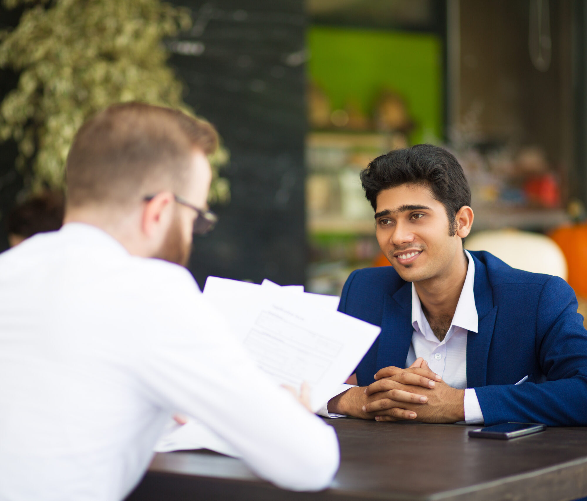 Smiling businessman waiting when partner reading contract in cafe. Content confident Indian manager sitting at table while his colleague analyzing papers. Examining documents concept