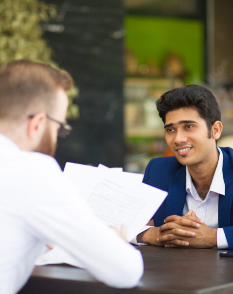 Smiling businessman waiting when partner reading contract in cafe. Content confident Indian manager sitting at table while his colleague analyzing papers. Examining documents concept